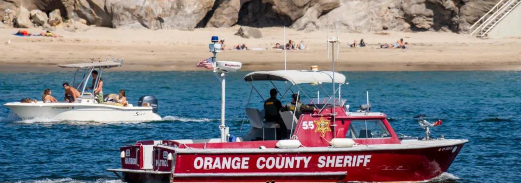 An Orange County Sheriff boat patrols the water near a beach with people sunbathing and another boat carrying passengers.