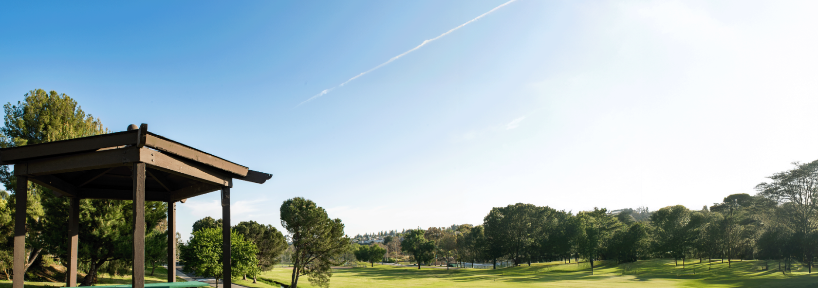 A picnic shelter with a table and benches sits on a paved area next to a lush green lawn and trees, with a golf course stretching into the distance under a bright blue sky with a faint contrail.