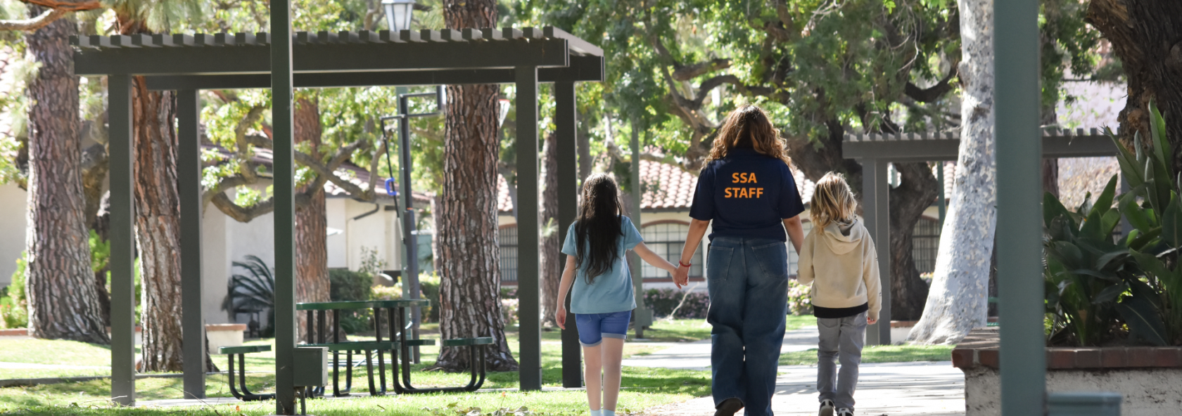 A person wearing an SSA STAFF shirt walks hand-in-hand with two children on a paved path through a park with trees and covered picnic areas.