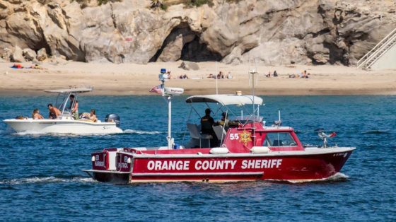 An Orange County Sheriff boat patrols the water near a beach with people sunbathing and another boat carrying passengers.