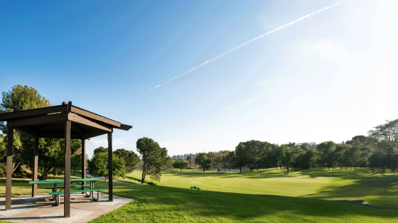 A picnic shelter with a table and benches sits on a paved area next to a lush green lawn and trees, with a golf course stretching into the distance under a bright blue sky with a faint contrail.