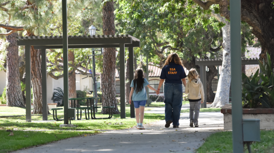 A person wearing an SSA STAFF shirt walks hand-in-hand with two children on a paved path through a park with trees and covered picnic areas.